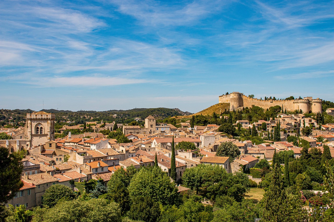 Panoramic view of a historic French village with terracotta rooftops and a medieval fortress on a hill under a bright blue sky.