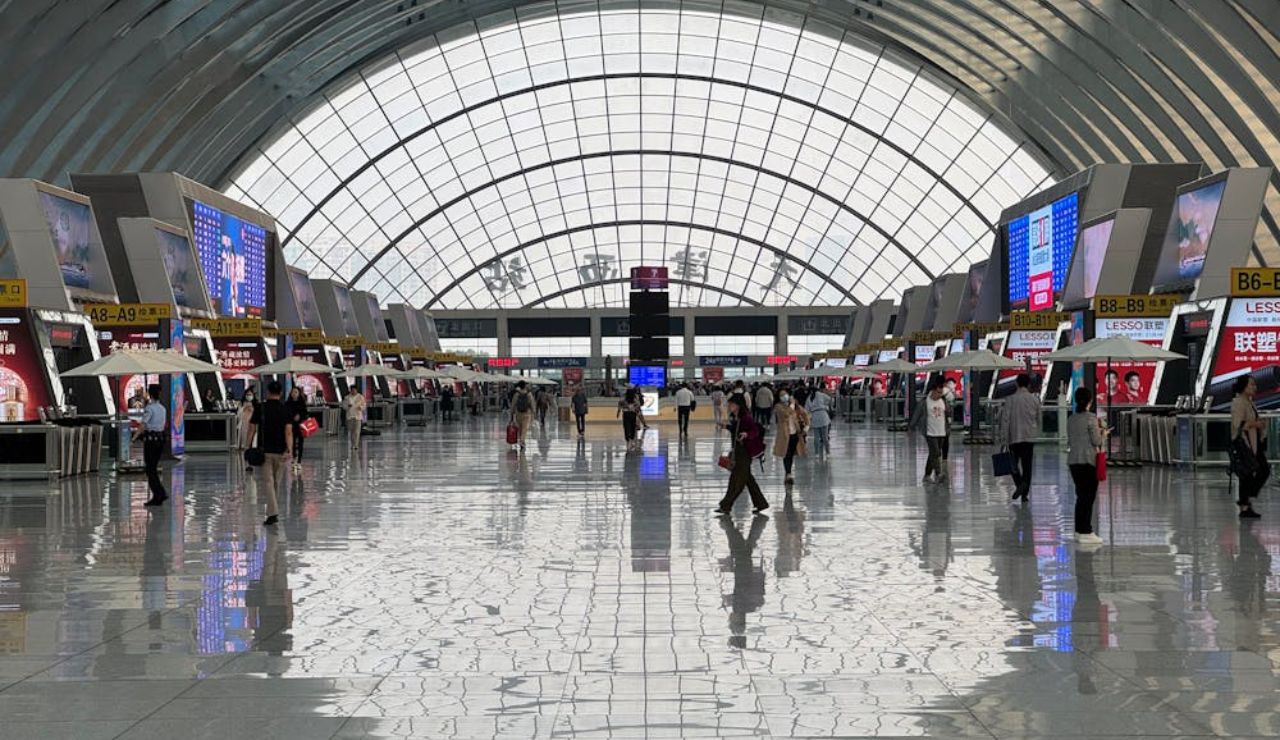 Wide interior view of a modern airport terminal with people walking and check-in counters on both sides under a large arched glass ceiling.