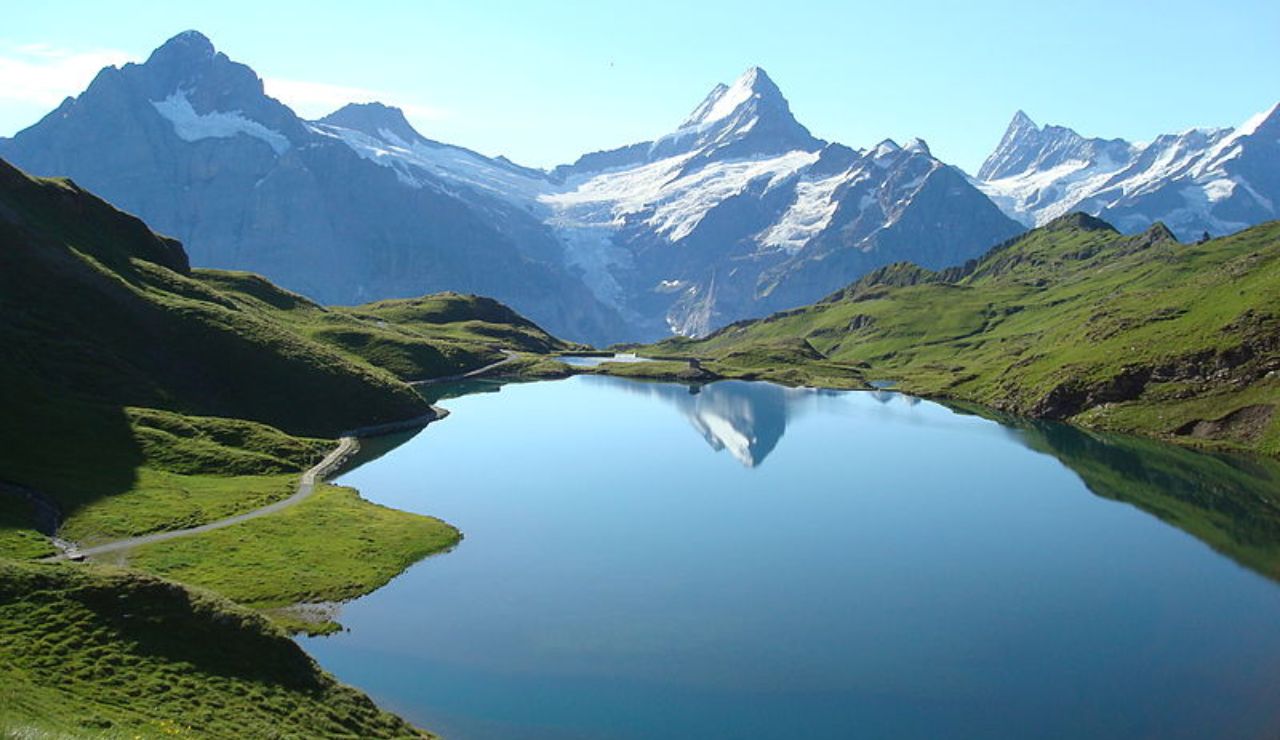Crystal-clear alpine lake reflecting snow-capped peaks and green hills, with a walking trail curving along the shoreline under a bright blue sky.