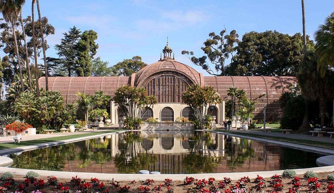 Historic botanical building with arched windows, surrounded by lush greenery and reflected in a large rectangular pond.