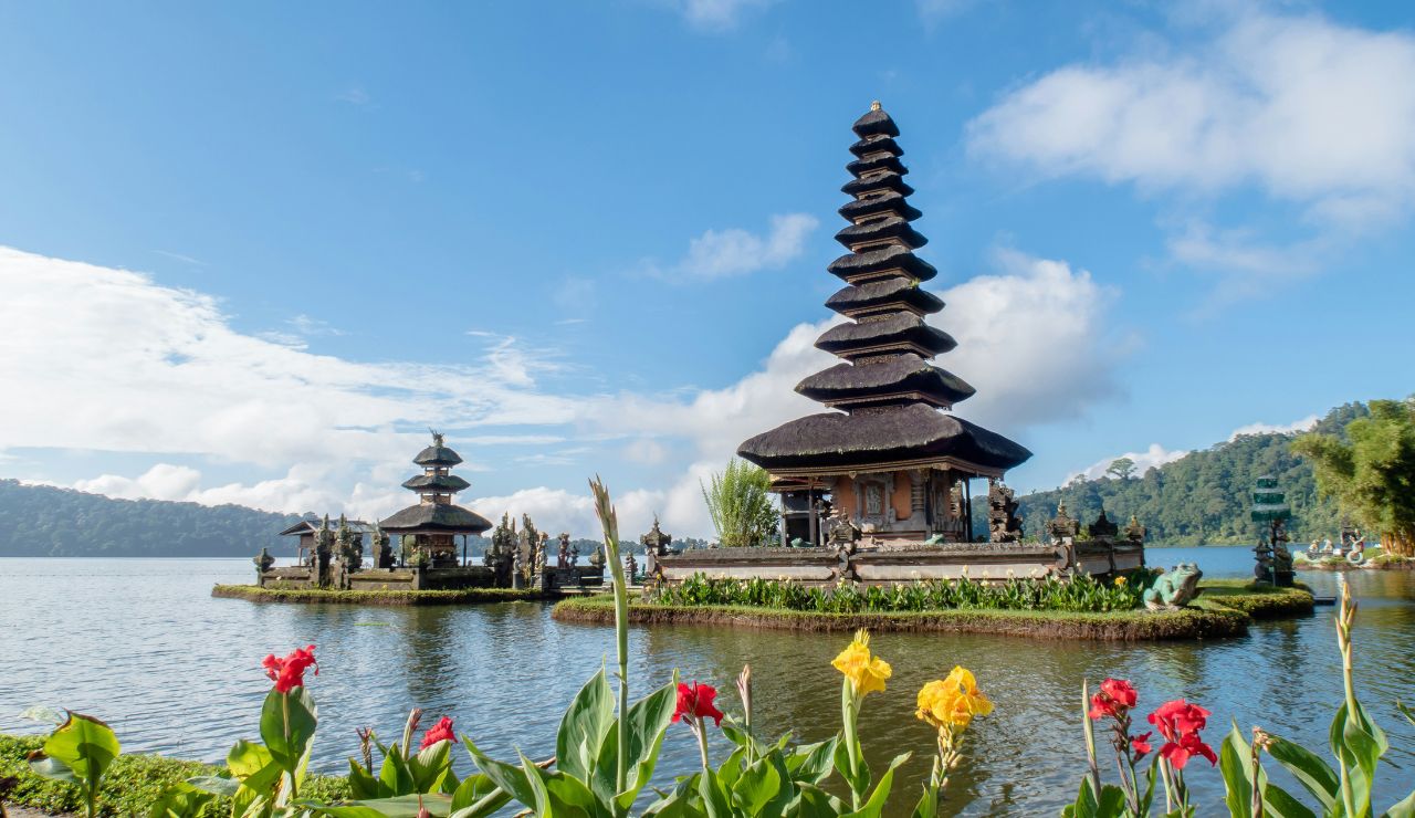 Traditional Balinese temple with multi-tiered roof sitting on a small island in a lake, surrounded by mountains and vibrant flowers in the foreground.