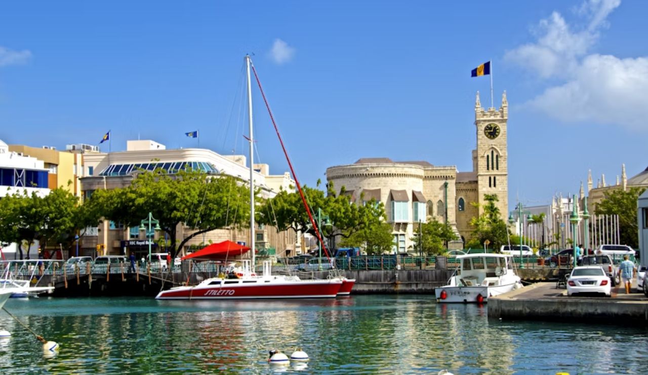 Harbor scene in Bridgetown, Barbados with boats docked on calm water and historic buildings including a clock tower flying the Barbados flag.