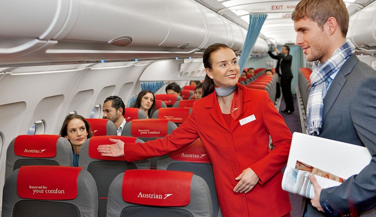 Austrian Airlines flight attendant in red uniform warmly guiding a male passenger to his seat inside the aircraft cabin.