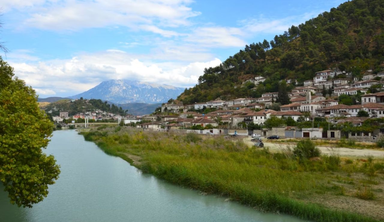 Riverside view of Berat, Albania, with traditional white Ottoman-style houses climbing up a green hillside and mountains in the background.