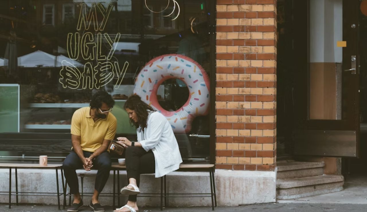 A couple sits on a bench outside a cafe with a large donut decoration and a neon sign reading "MY UGLY BABY," casually chatting and looking at a phone.