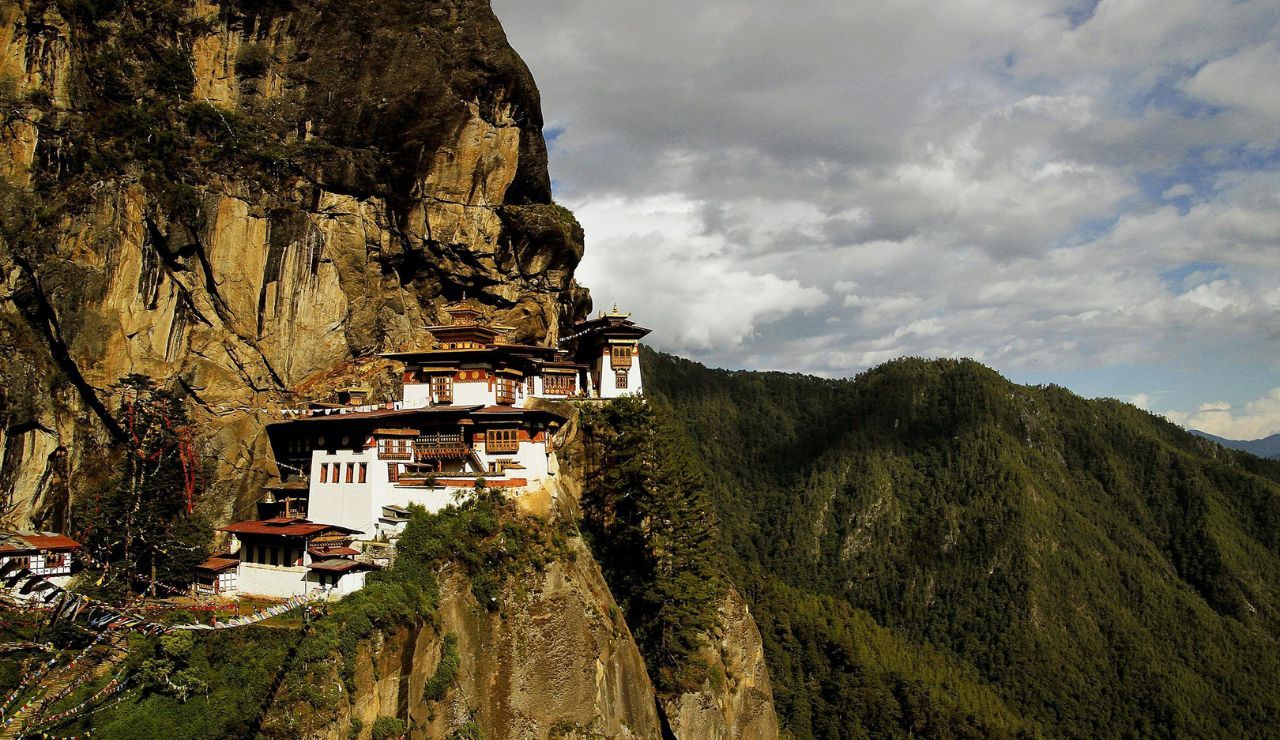 Monastery perched dramatically on a cliffside overlooking lush green mountains under a partly cloudy sky.