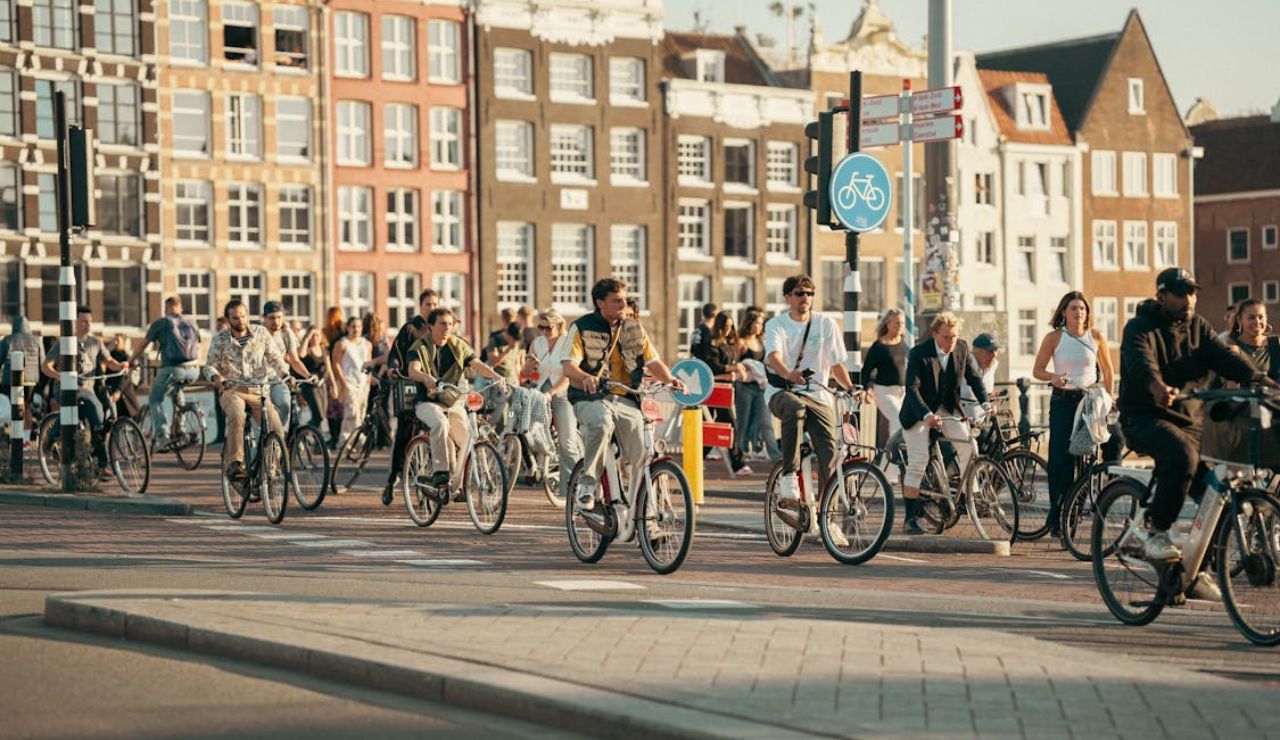 A large group of cyclists ride through a busy bike lane in a European city, surrounded by pedestrians and historic buildings in the background.