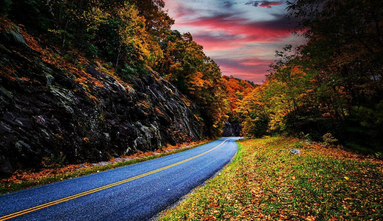 Scenic autumn road winding through rocky hills and vibrant fall foliage under a dramatic red and purple sunset sky.