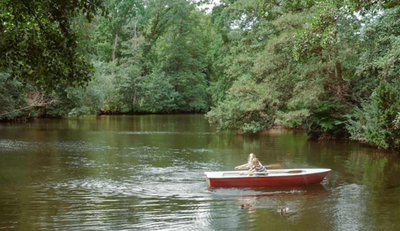 Woman rowing a small red and white boat on a calm forested lake surrounded by lush green trees.