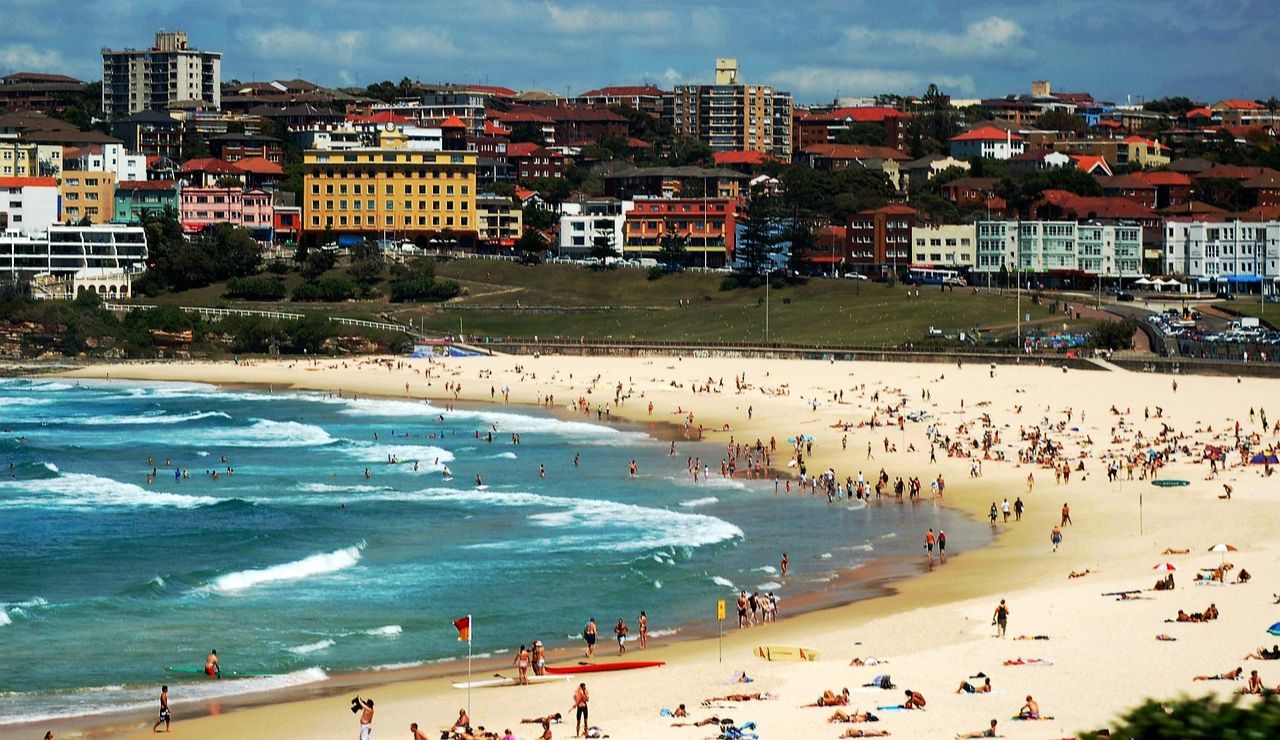 Crowded sandy beach with people swimming and sunbathing, set against a backdrop of colorful coastal buildings and urban skyline.