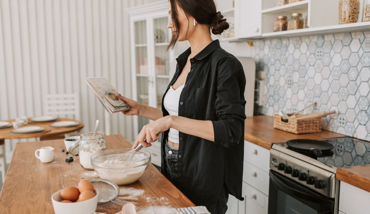 Woman in a kitchen reading a cookbook while whisking batter in a bowl.