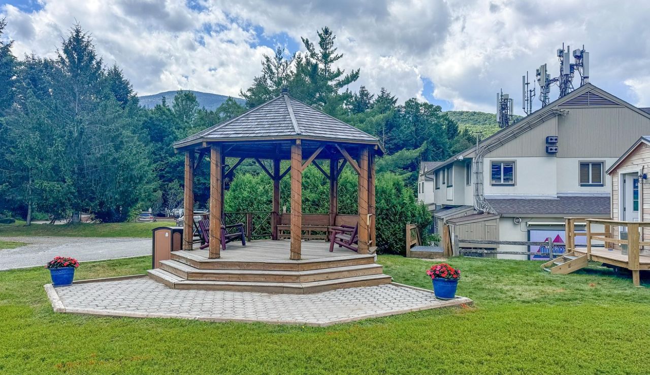 Wooden gazebo on a paved platform with benches, surrounded by greenery and buildings in the background.