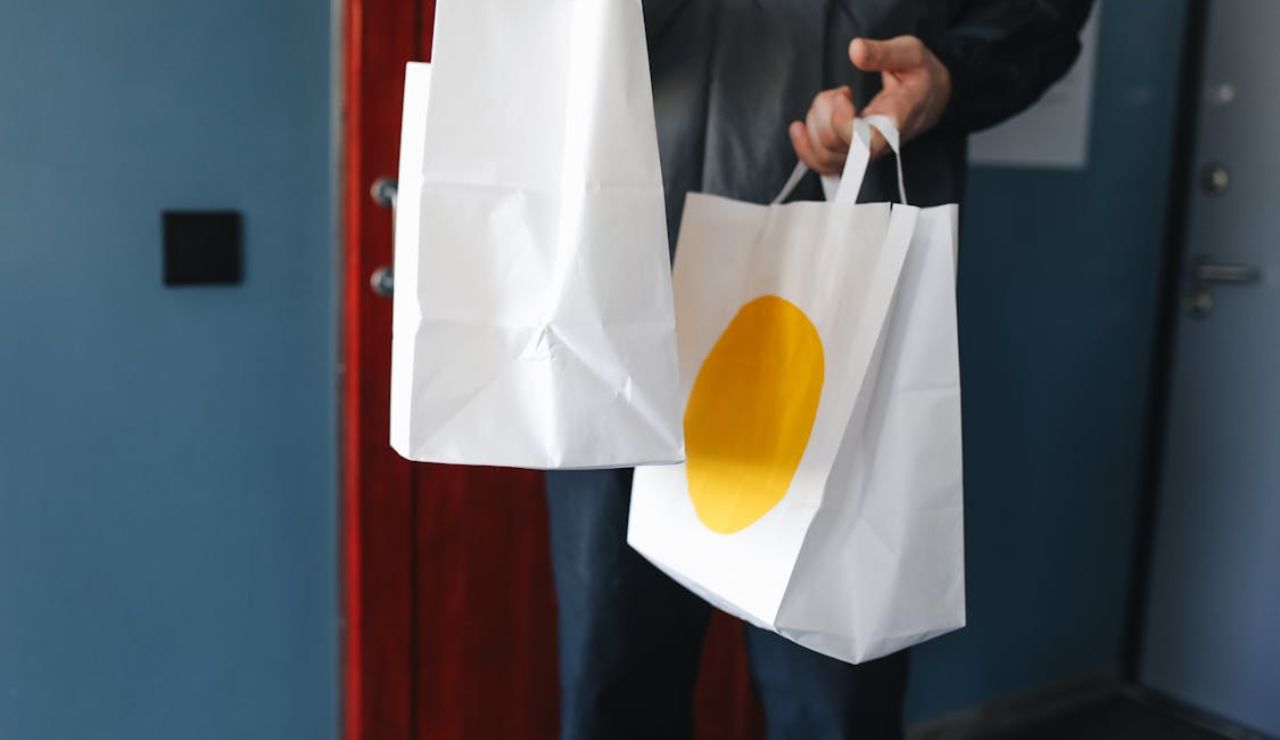 Person holding two white paper bags, one with a yellow circle, standing near a doorway.