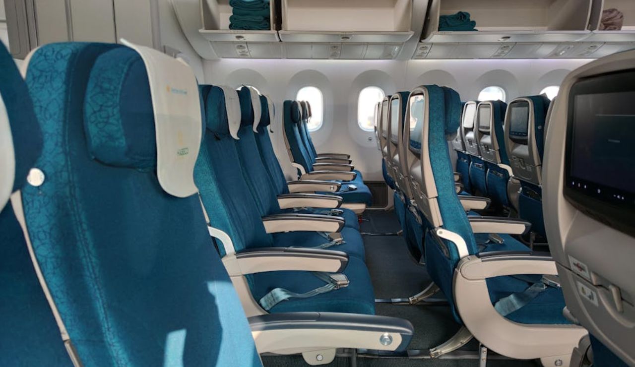 Empty airplane cabin with rows of blue economy seats, seatbelts neatly arranged, and blankets stored in overhead compartments.