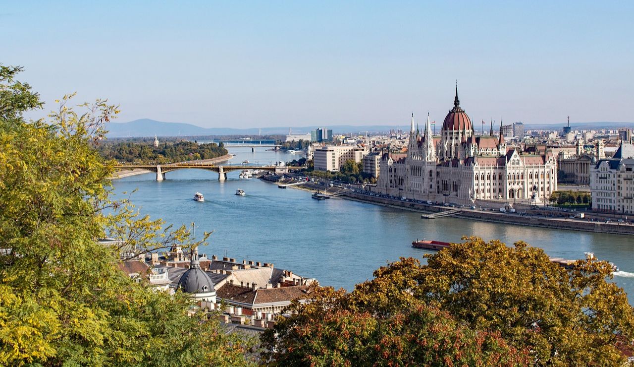 Panoramic view of Budapest with the Hungarian Parliament building along the Danube River and boats sailing under a bridge.