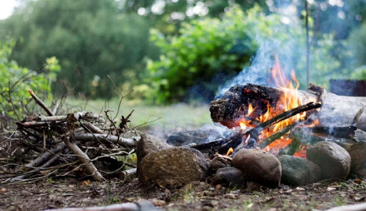 Campfire burning inside a stone ring with logs and smoke rising, surrounded by forest greenery and camping tools.