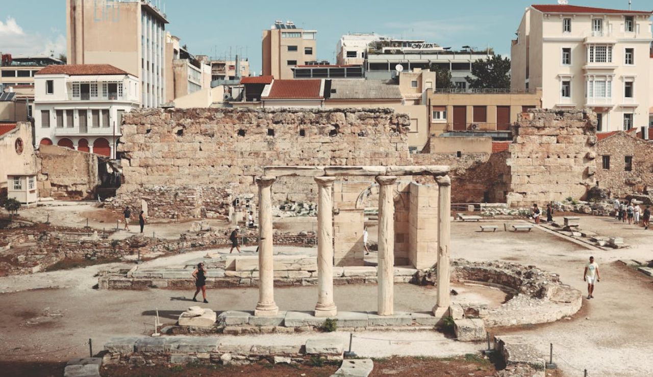 Ancient ruins with standing columns in front of a large stone wall, surrounded by modern buildings in a city setting.