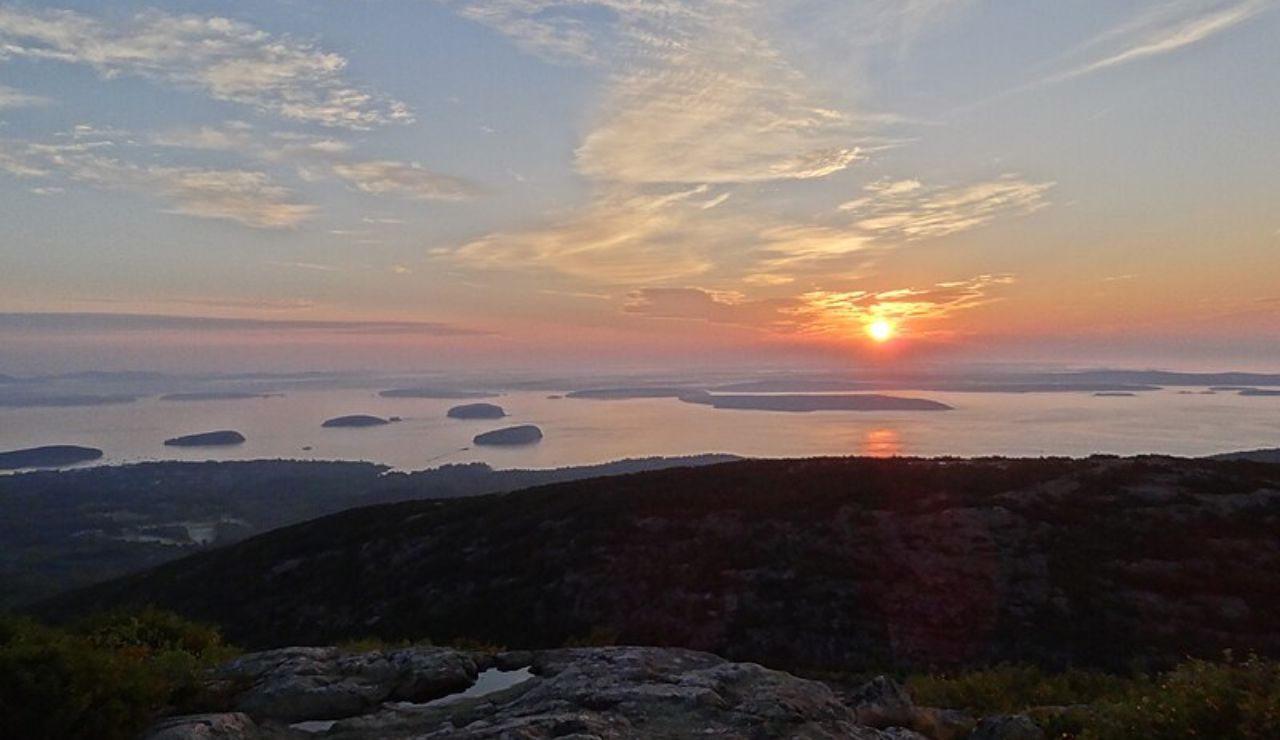 Sunrise over the ocean as seen from a mountain viewpoint, with scattered islands and soft clouds reflecting warm morning light.
