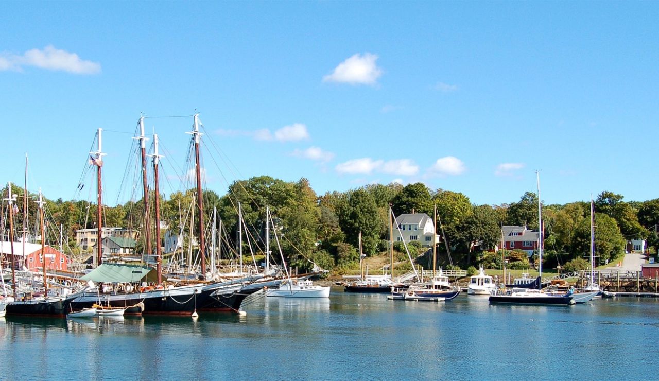 Harbor with sailboats and historic ships docked near a quaint coastal town on a sunny day.