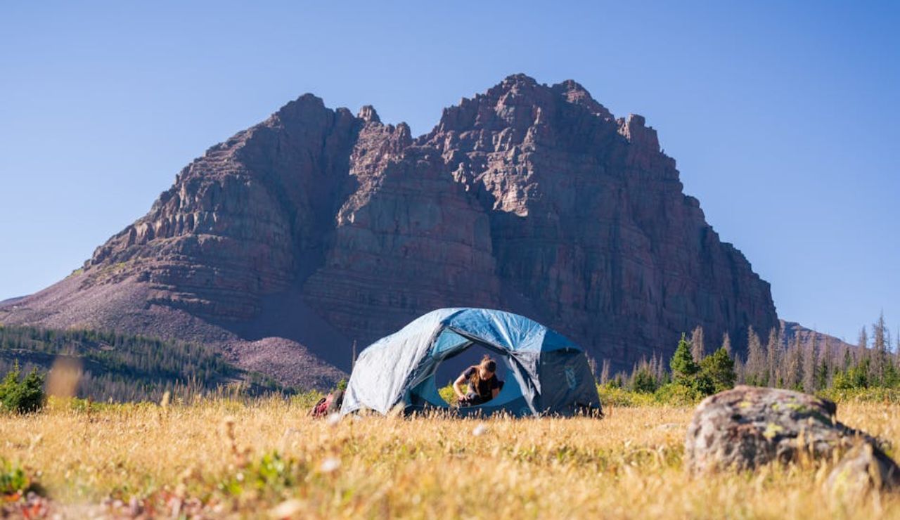 Person inside a blue tent on a grassy field with a large rocky mountain in the background under a clear blue sky.