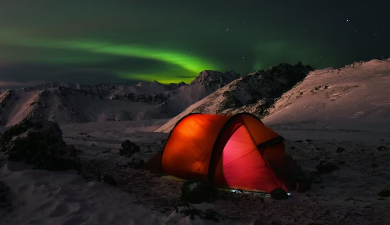 Glowing orange tent in snowy mountains under the Northern Lights at night.