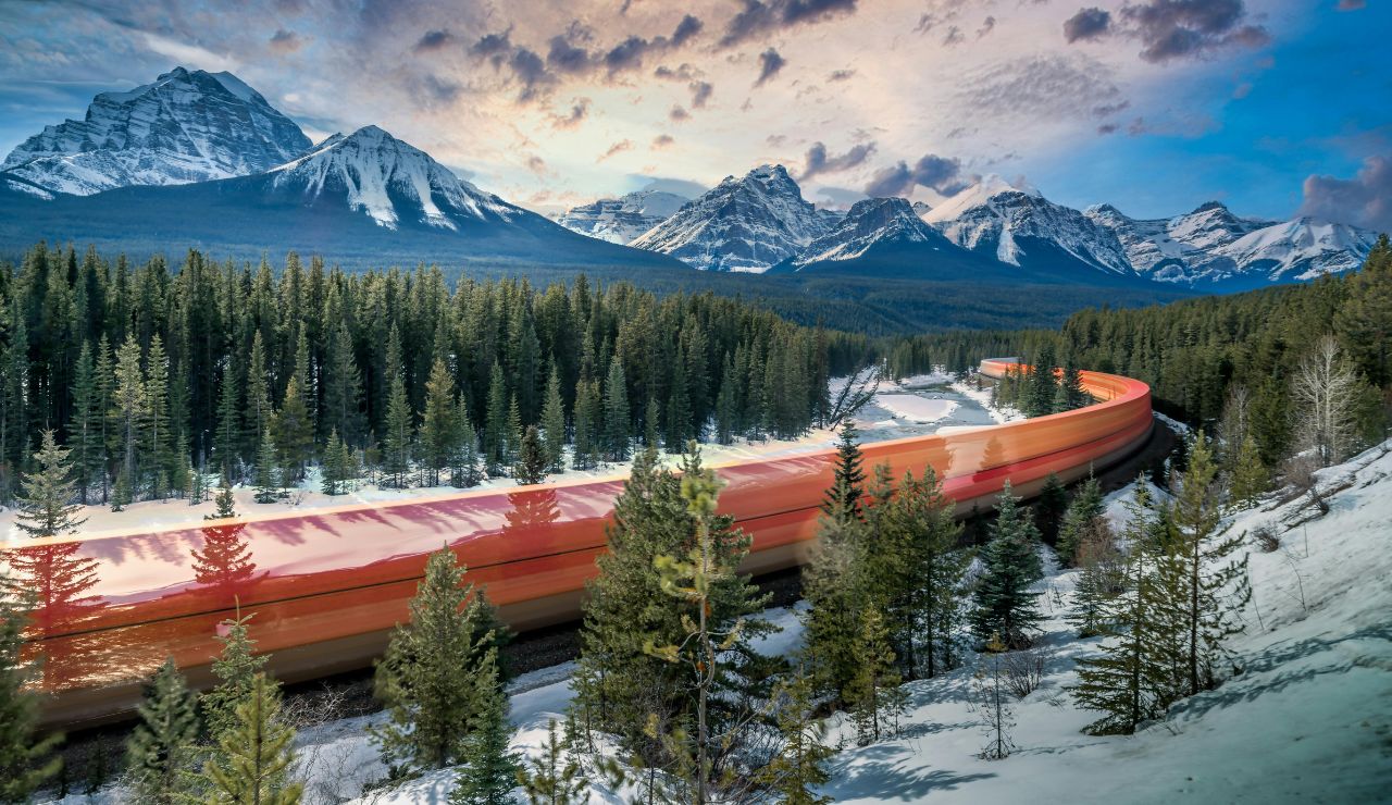 A passenger train winding through forested mountains and lakes in Canada.