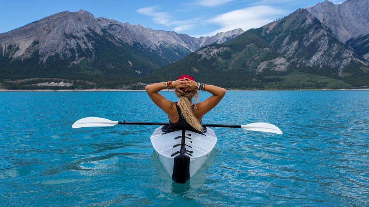 Woman kayaking on a turquoise lake surrounded by mountains, relaxing with hands behind her head.