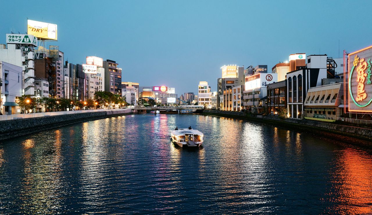 A boat travels down a river past illuminated city buildings and signs at dusk.