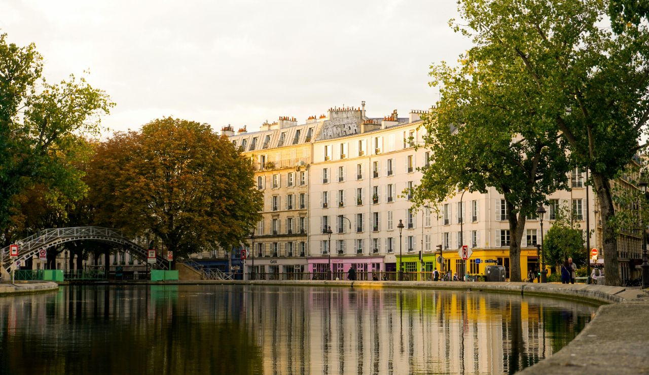 A calm canal curves past trees and colorful storefronts beneath multistory buildings, with a metal footbridge crossing on the left under a partly cloudy sky.