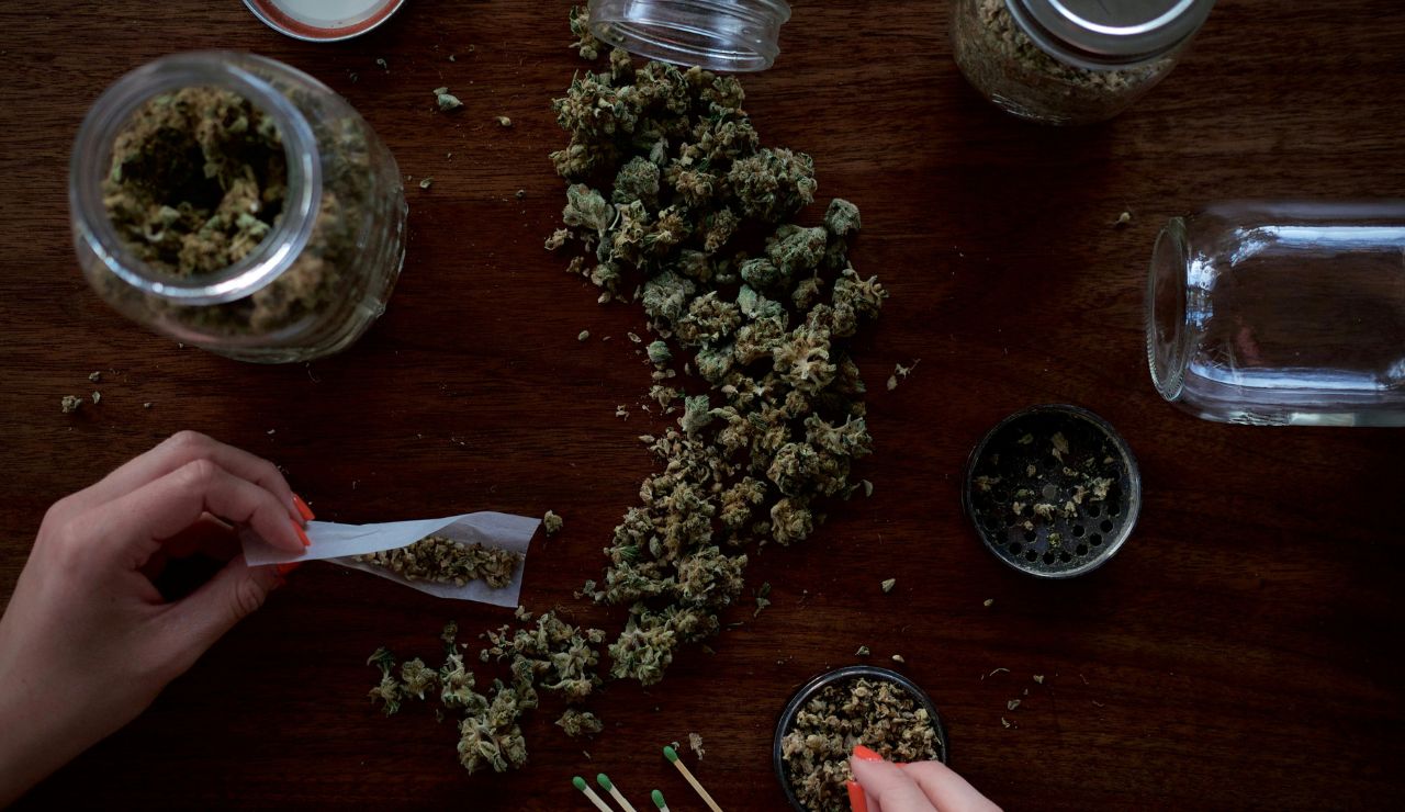 Person rolling a joint with cannabis buds and jars on a wooden table.