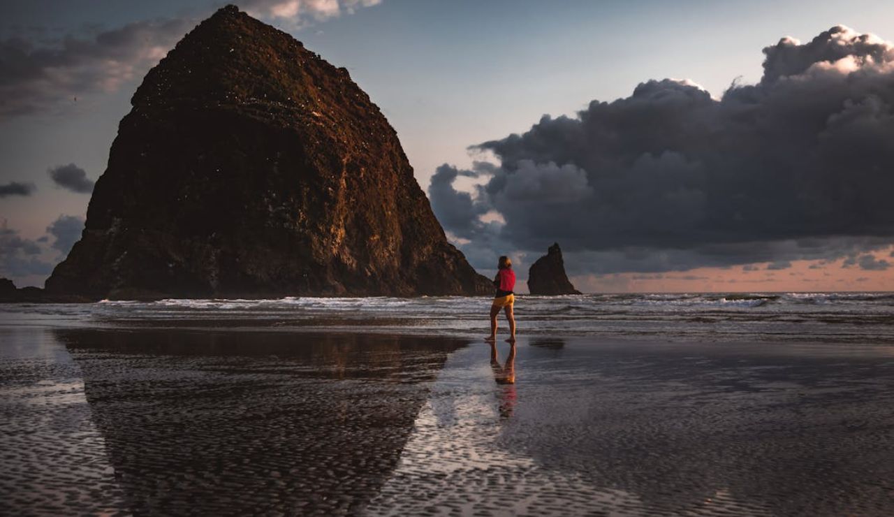 Person walking along the wet sand at Cannon Beach, Oregon, with Haystack Rock towering in the background under dramatic evening clouds.