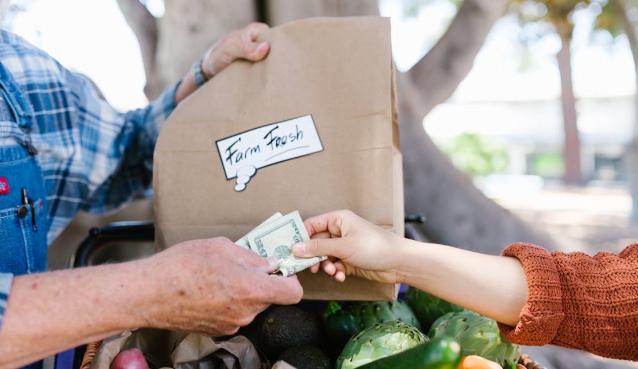Customer handing cash to a vendor for a paper bag labeled "Farm Fresh" at a produce stand.
