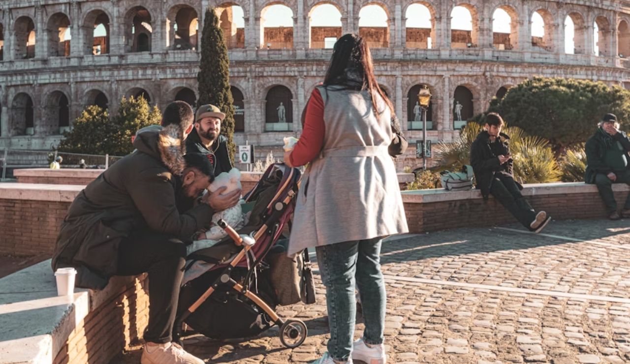 A family near the Colosseum, with one person sitting on a bench holding a baby, while others are standing or sitting nearby, enjoying the scene.