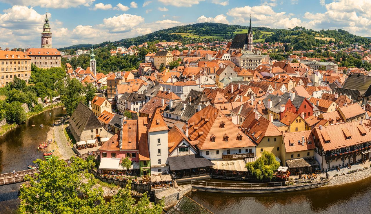 Panoramic view of a European town with red-roofed buildings, a river, and green hills beneath a partly cloudy sky.