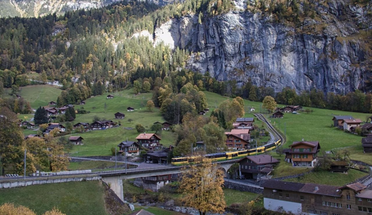 A train passing through a picturesque Swiss village, surrounded by lush green fields and towering cliffs.