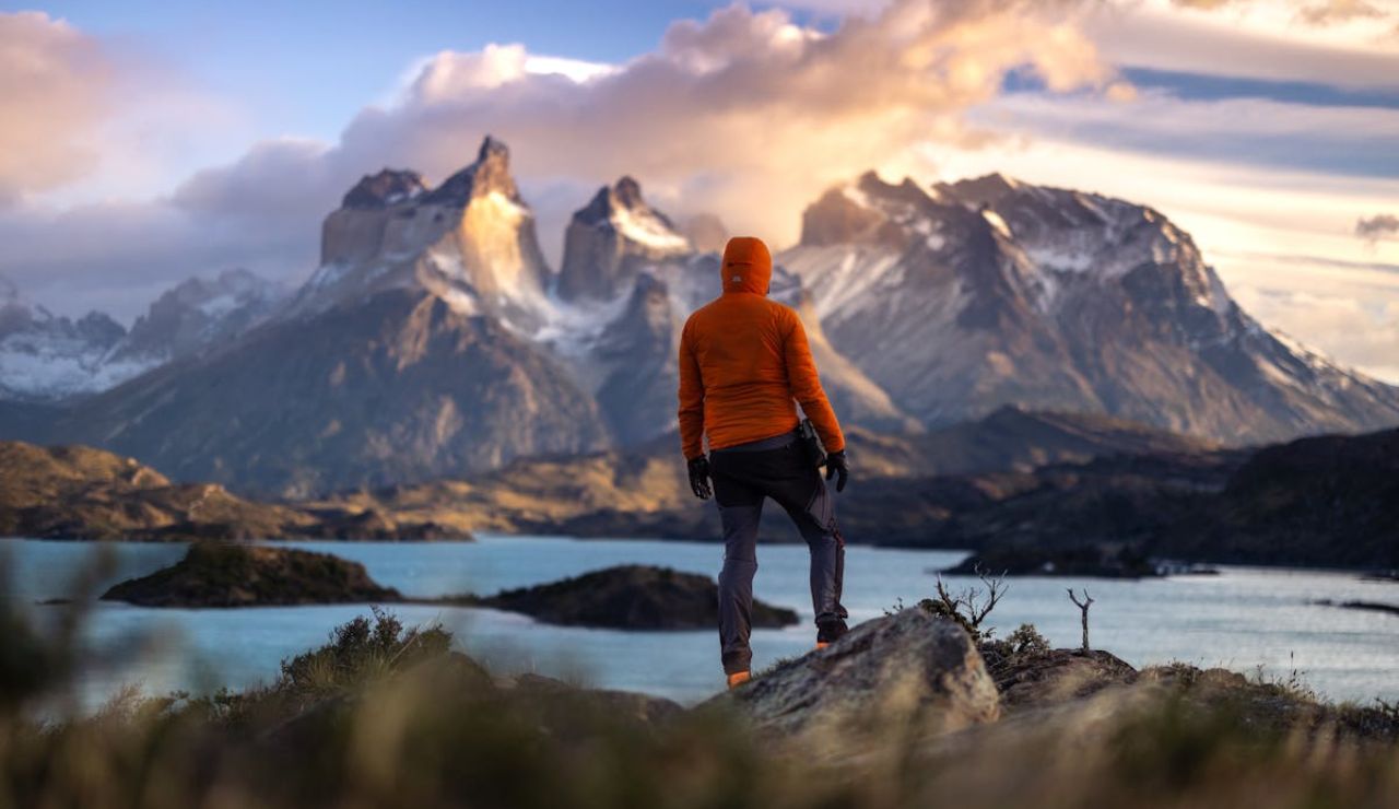 Hiker in an orange jacket overlooking dramatic mountain peaks and turquoise lakes at sunset.