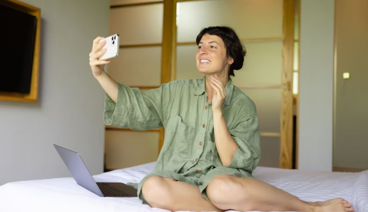 Woman sitting on bed in casual clothes, smiling and waving while taking a selfie with a smartphone; an open laptop rests beside her.