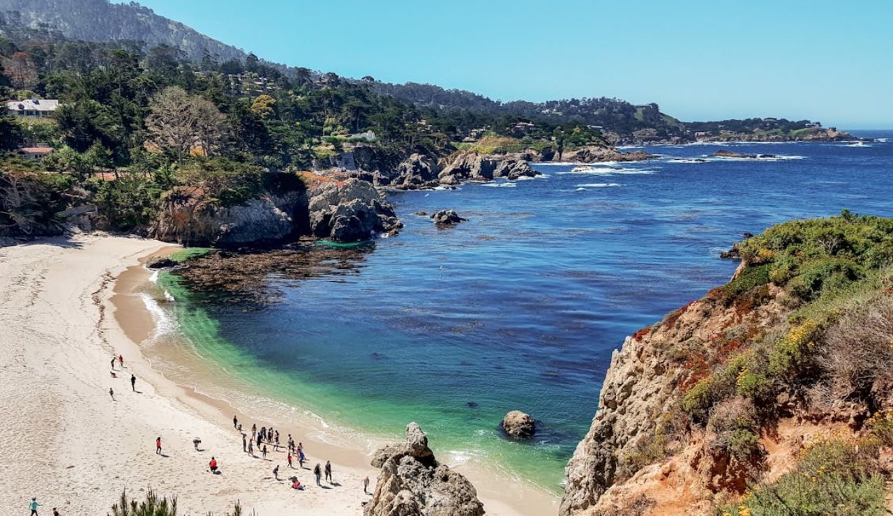  Scenic coastal beach with clear blue water, rocky cliffs, and people walking on the sand.