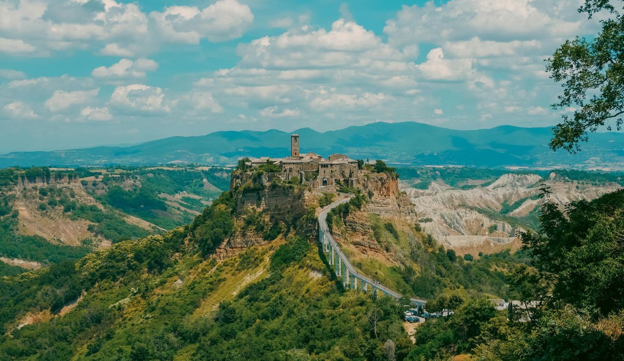 A hilltop village of stone buildings and a tall central tower is linked to the land by a narrow bridge, set against mountains and greenery.