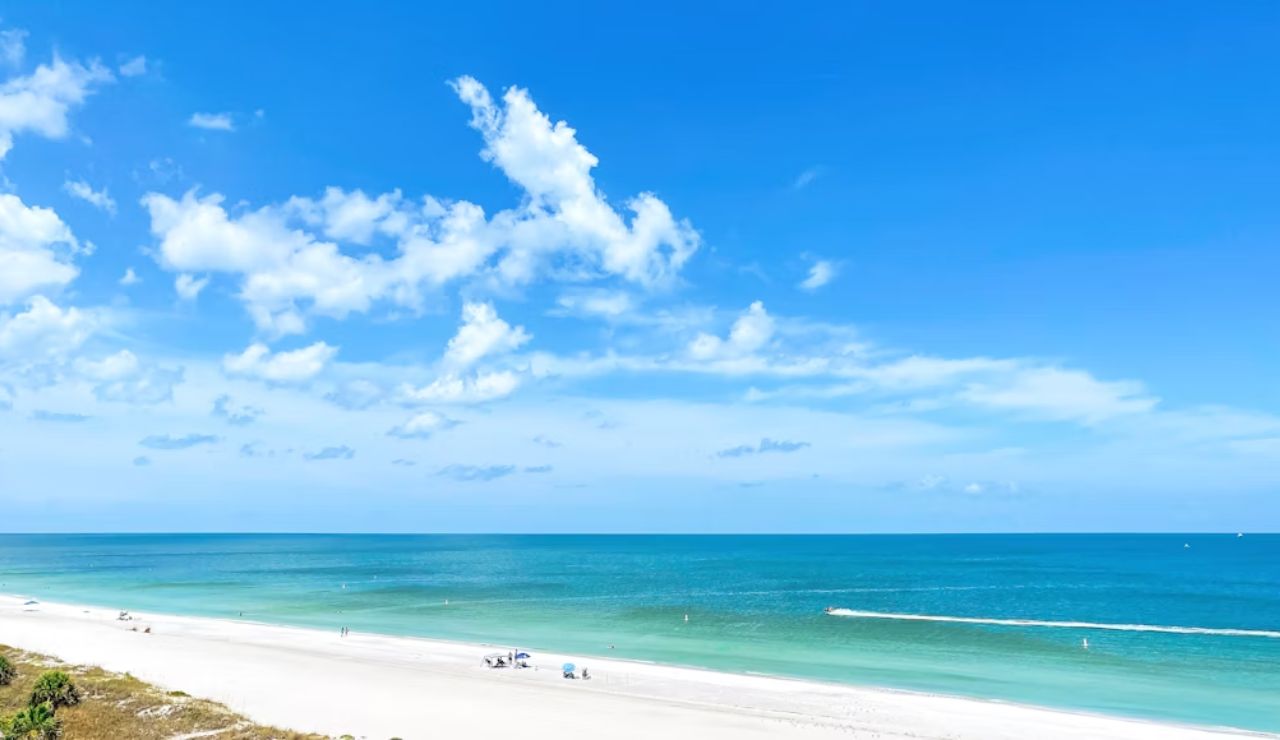 A scenic view of a pristine white sand beach with turquoise water and a bright blue sky dotted with clouds.