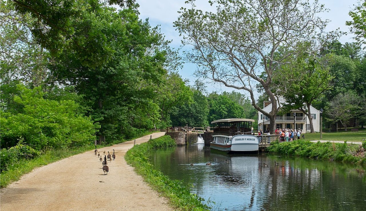 Scenic view of the C&O Canal Towpath with ducks on the trail, a historic canal boat on the water, and visitors gathered near a white house.
