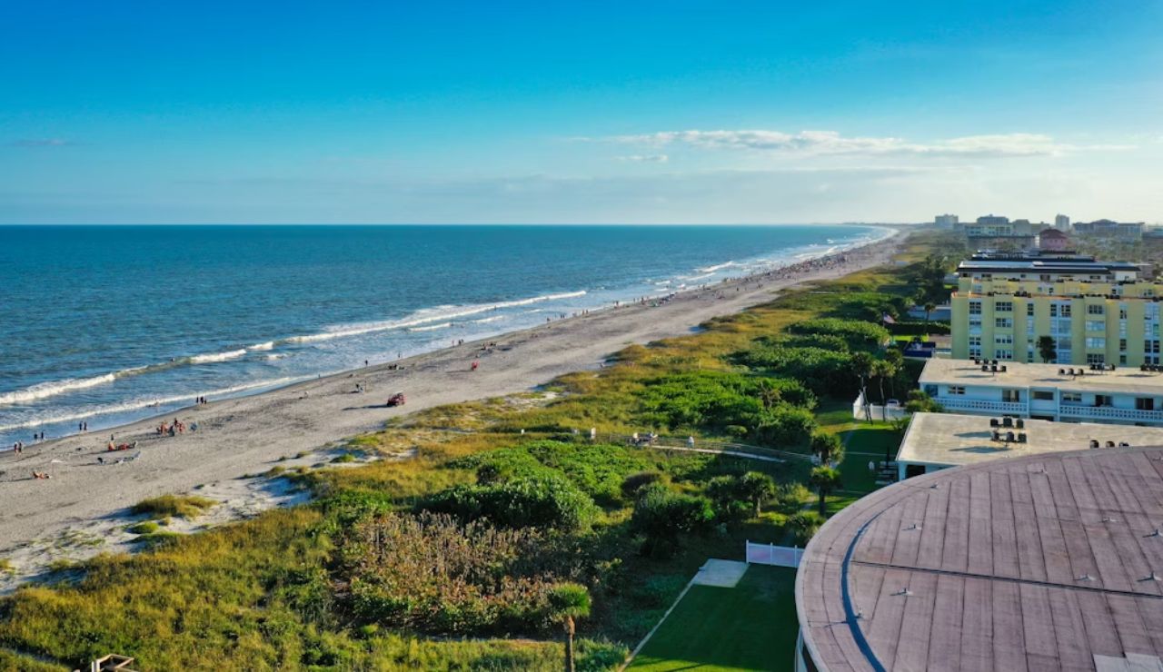 A coastal view of a beach lined with buildings and greenery, with waves gently rolling in under a clear blue sky.
