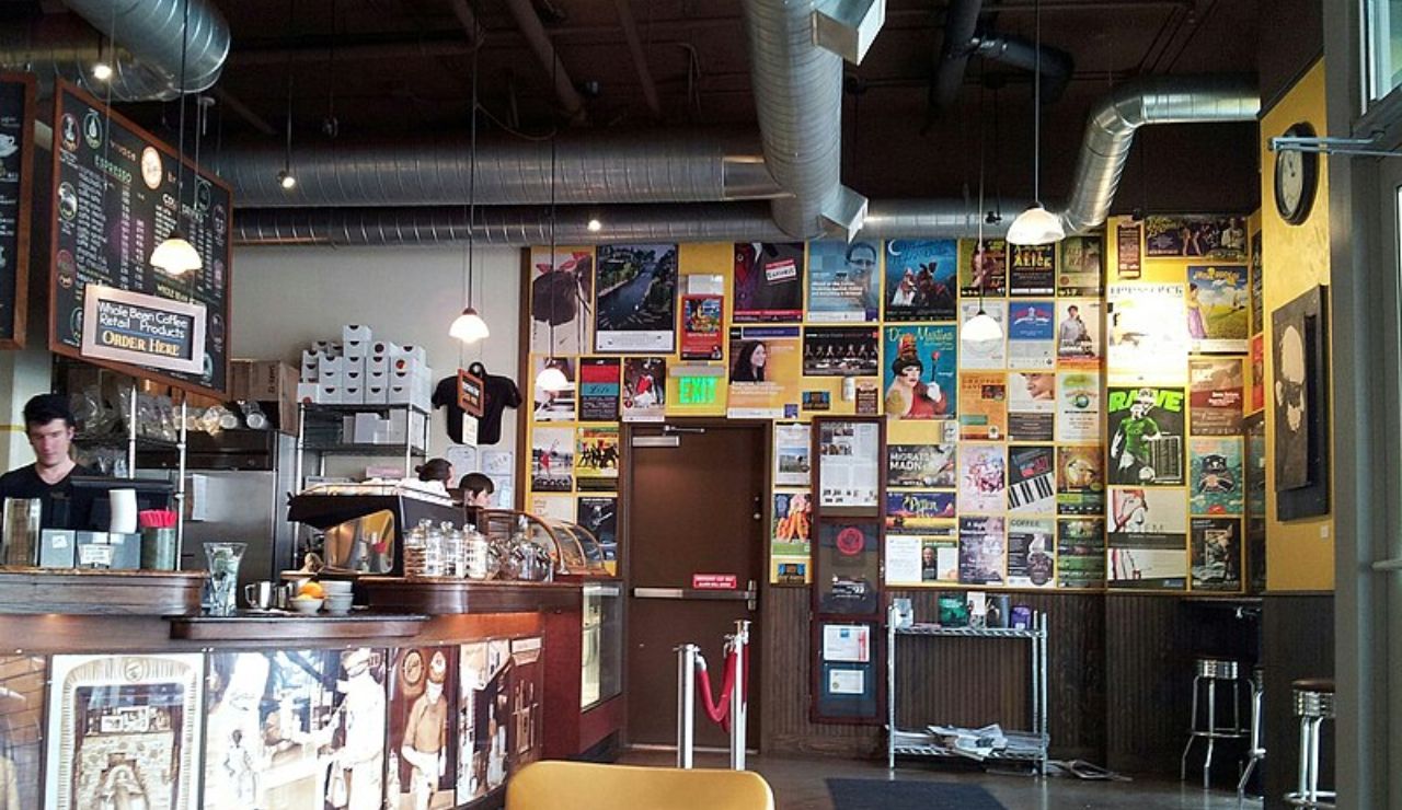 A cozy café interior with a busy coffee counter, industrial decor, and a wall filled with colorful posters.