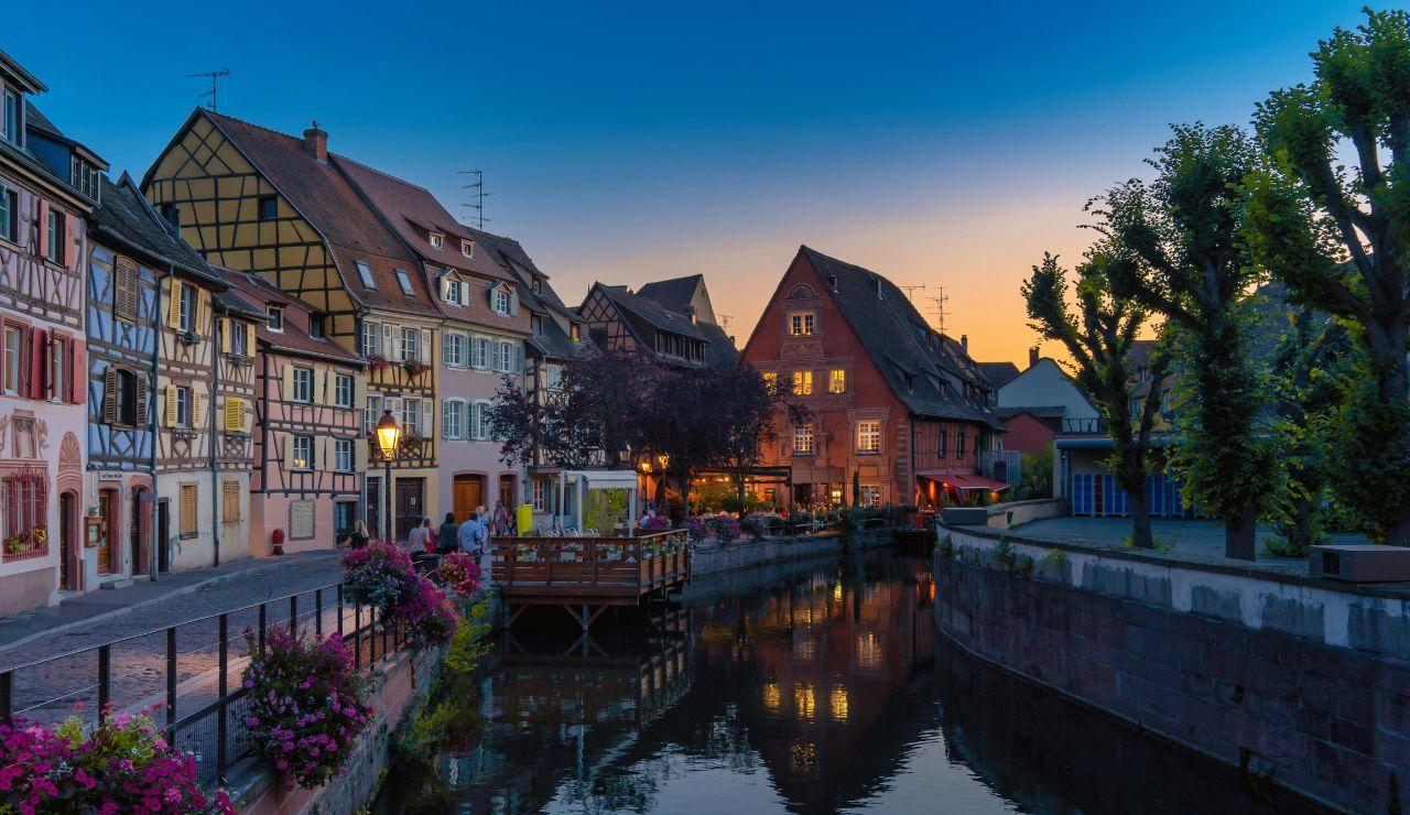 Colorful traditional European houses with flower boxes line a calm canal at sunset, while people walk and sit outside among trees.