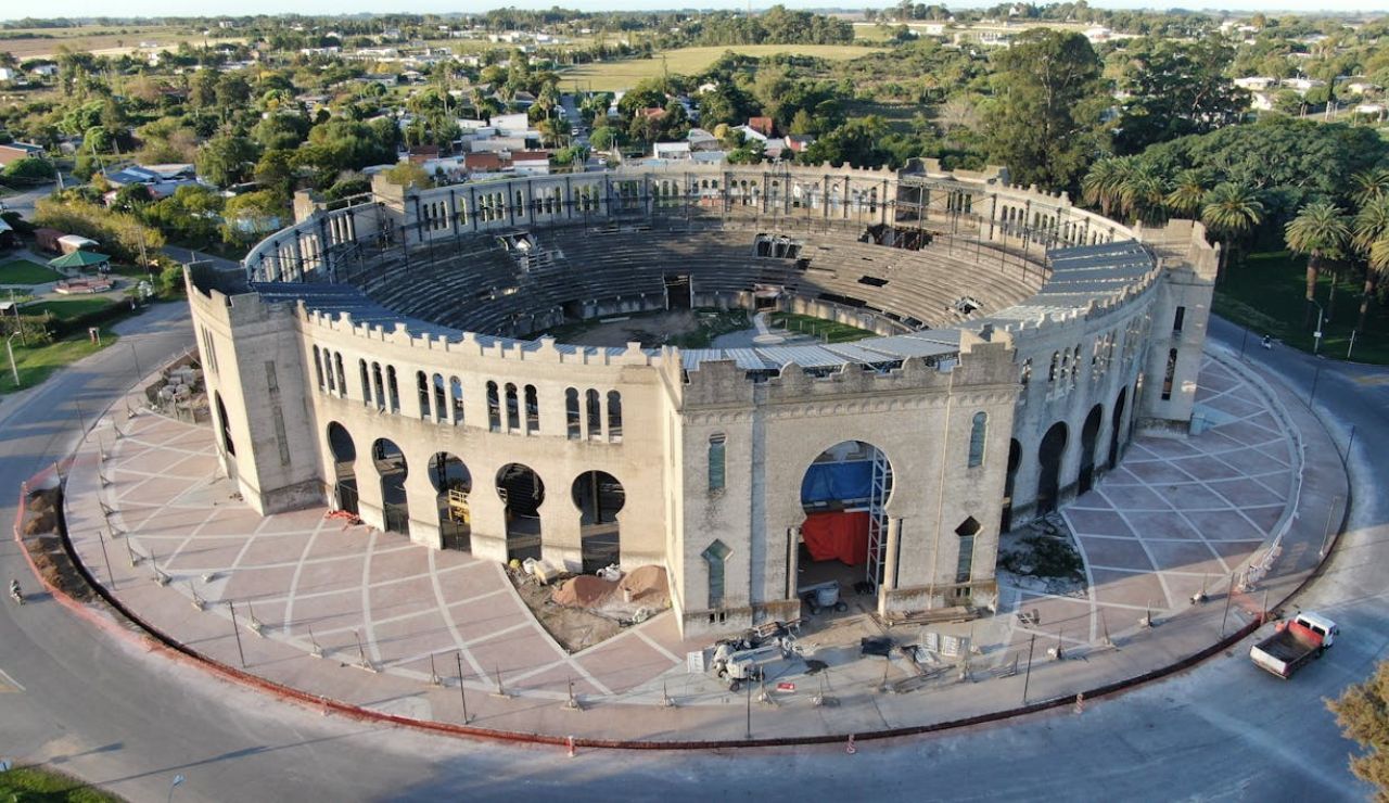 Aerial view of an old stone bullring with an open circular arena, surrounded by a paved plaza and green suburban landscape.