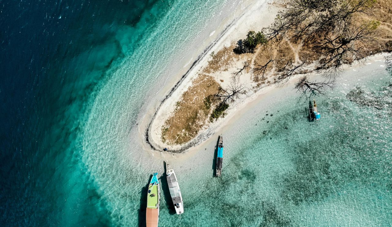 Aerial view of a sandy peninsula with three boats anchored in clear turquoise water near the shore.