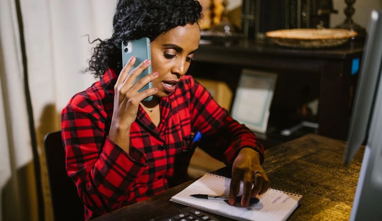 Woman in red plaid shirt talking on phone while taking notes at a desk.