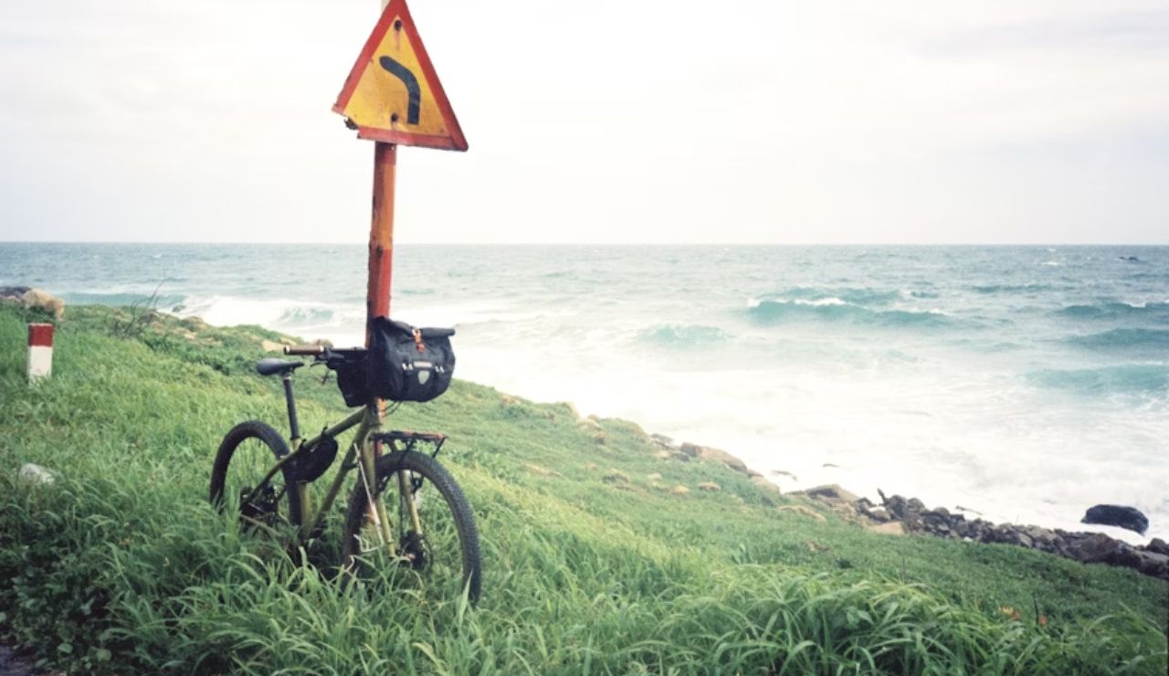Bicycle parked against a left-turn road sign on a grassy coastal path beside the ocean.