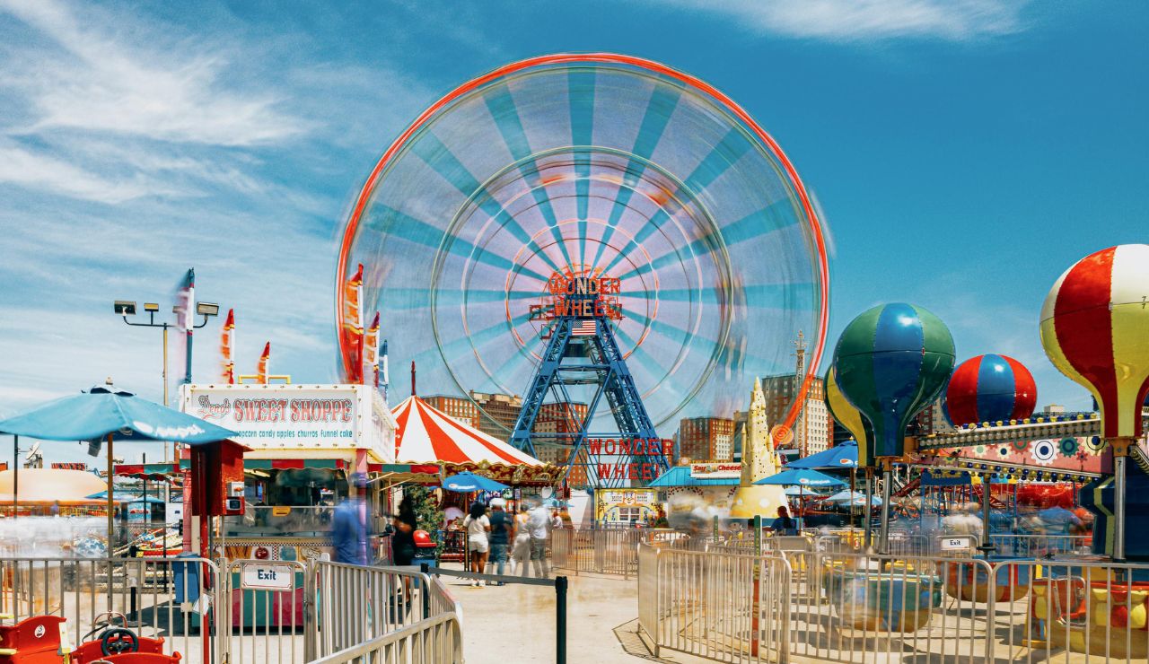 People enjoying a sunny day at Coney Island amusement park with the Wonder Wheel spinning in the background and colorful rides in the foreground.