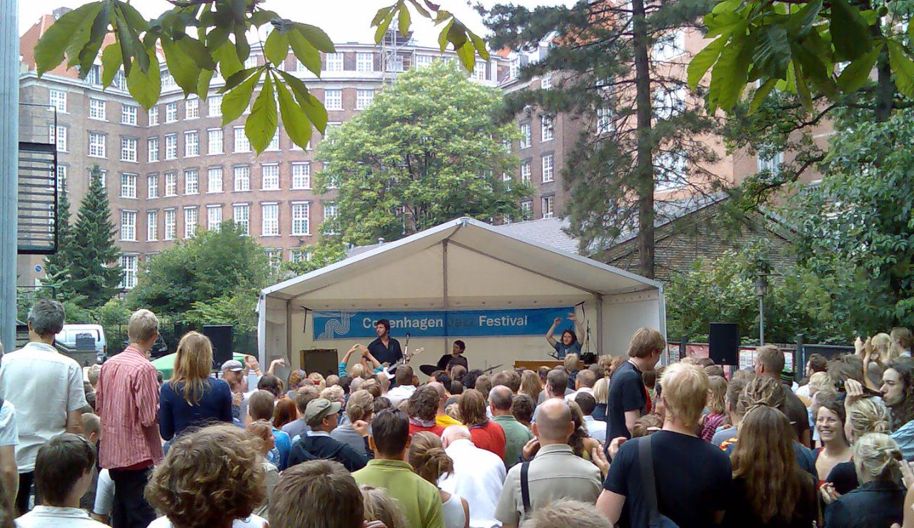 A crowd gathers in a park for a live outdoor performance during the Copenhagen Jazz Festival, with musicians playing on a small covered stage.